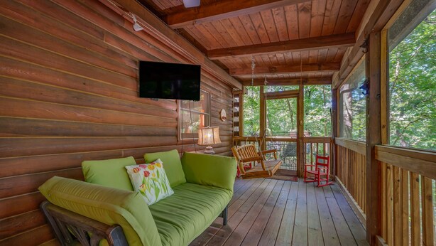 Een houten chalet met een groene bank, een televisie en een schommelstoel op een veranda met uitzicht op bomen.