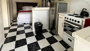 Una habitación de hotel pequeña con piso a cuadros, una cama, una kitchenette con estufa y un ventilador de techo.