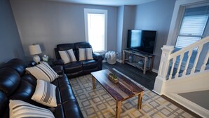 A living room with a black leather sofa, a wooden coffee table, a TV on a stand, and a staircase with white railings.