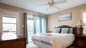 Une chambre à coucher comprenant un lit, un bureau, une chaise, un ventilateur de plafond et une fenêtre avec des rideaux.