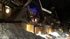 A snow-covered pathway leading to traditional wooden buildings with illuminated windows and a blue-lit structure in the background.
