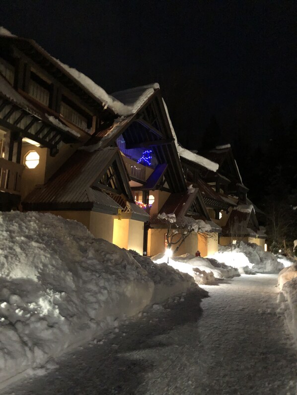 Un sentier enneigé mène à des bâtiments traditionnels en bois, dont les fenêtres sont illuminées, et une structure éclairée de lumière bleue se détache à l’arrière-plan.