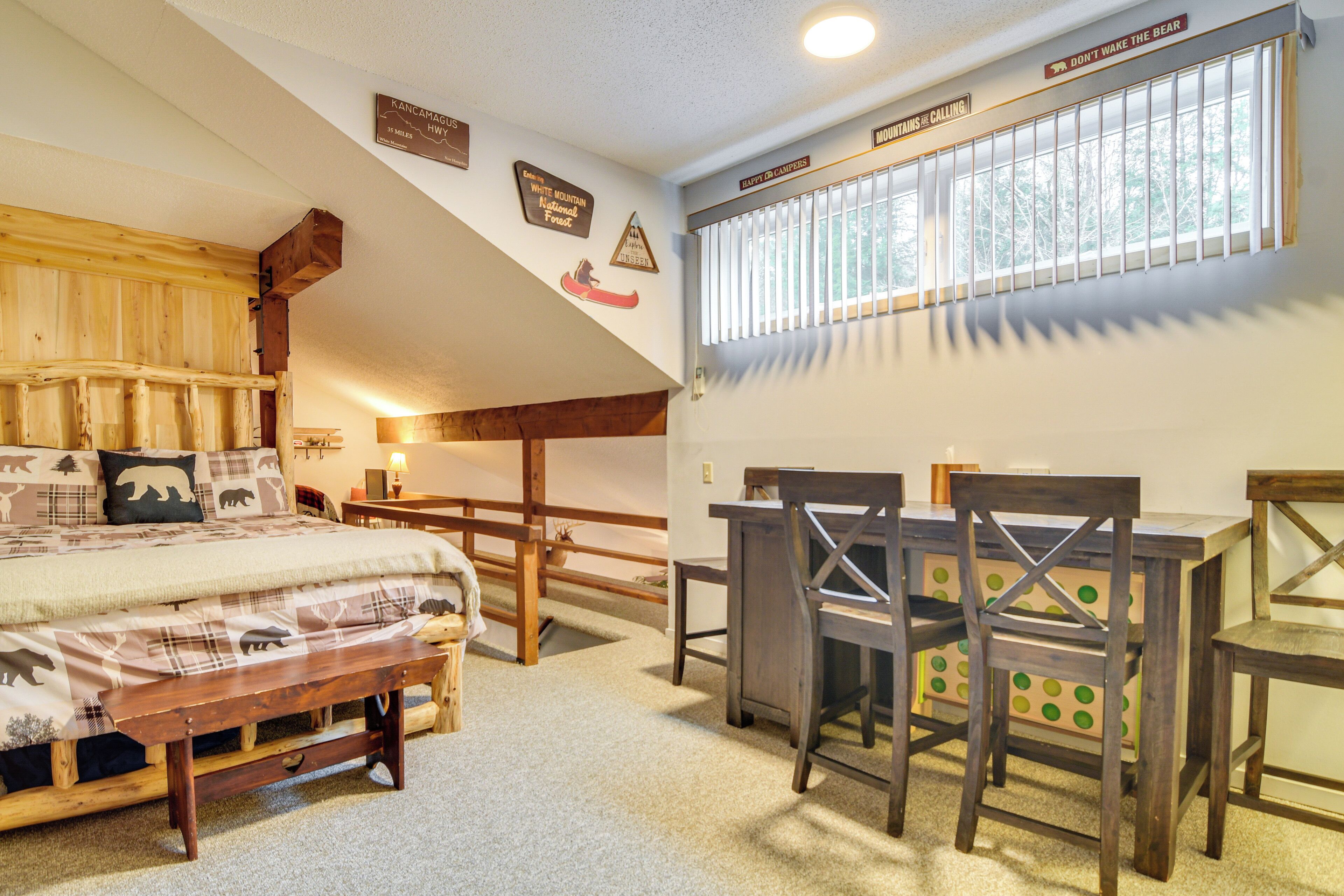 Una habitación acogedora con cama, un banco de madera y una mesa con sillas. Se observan adornos en las paredes y una ventana con persianas.