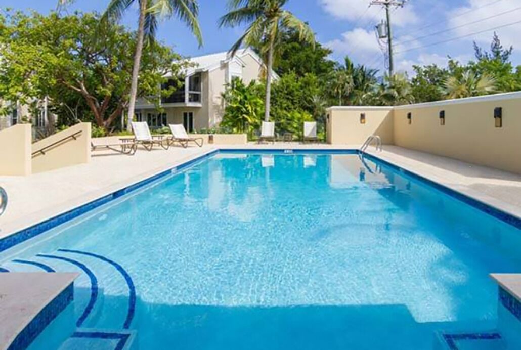 Une piscine rectangulaire à l’eau d’un bleu limpide, entourée d’une zone de bord de piscine de couleur beige, de palmiers et d’un bâtiment à la façade blanche en arrière-plan.