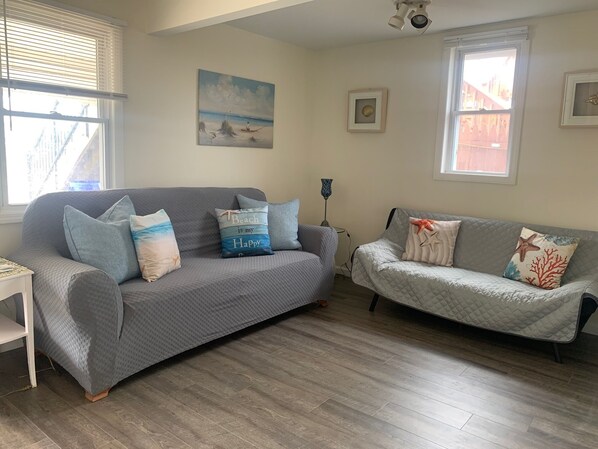 A living room with two grey sofas, one with a beach-themed pillow and the other with starfish pillows. There is a painting of a beach scene on the wall, a small table with a lamp, and a window with blinds.