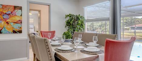 A dining area with a wooden table set for six, surrounded by upholstered chairs. A large potted plant is in the center, and a colorful abstract painting is on the wall.