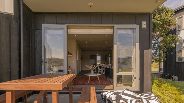 Een open terras met een houten tafel en bankjes, een zicht op het interieur door glazen deuren, en een klein zitgedeelte met een gestreept kussen.