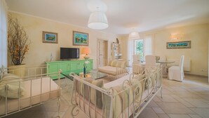 A living room with a sofa, a television, a lamp, and a dining table. - Villa Capo Perla immersed in a magnolia garden a few steps from the sea (Capoliveri)
