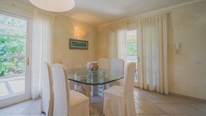 A dining area with a glass table, white chairs, and a painting on the wall. - Villa Capo Perla immersed in a magnolia garden a few steps from the sea (Capoliveri)