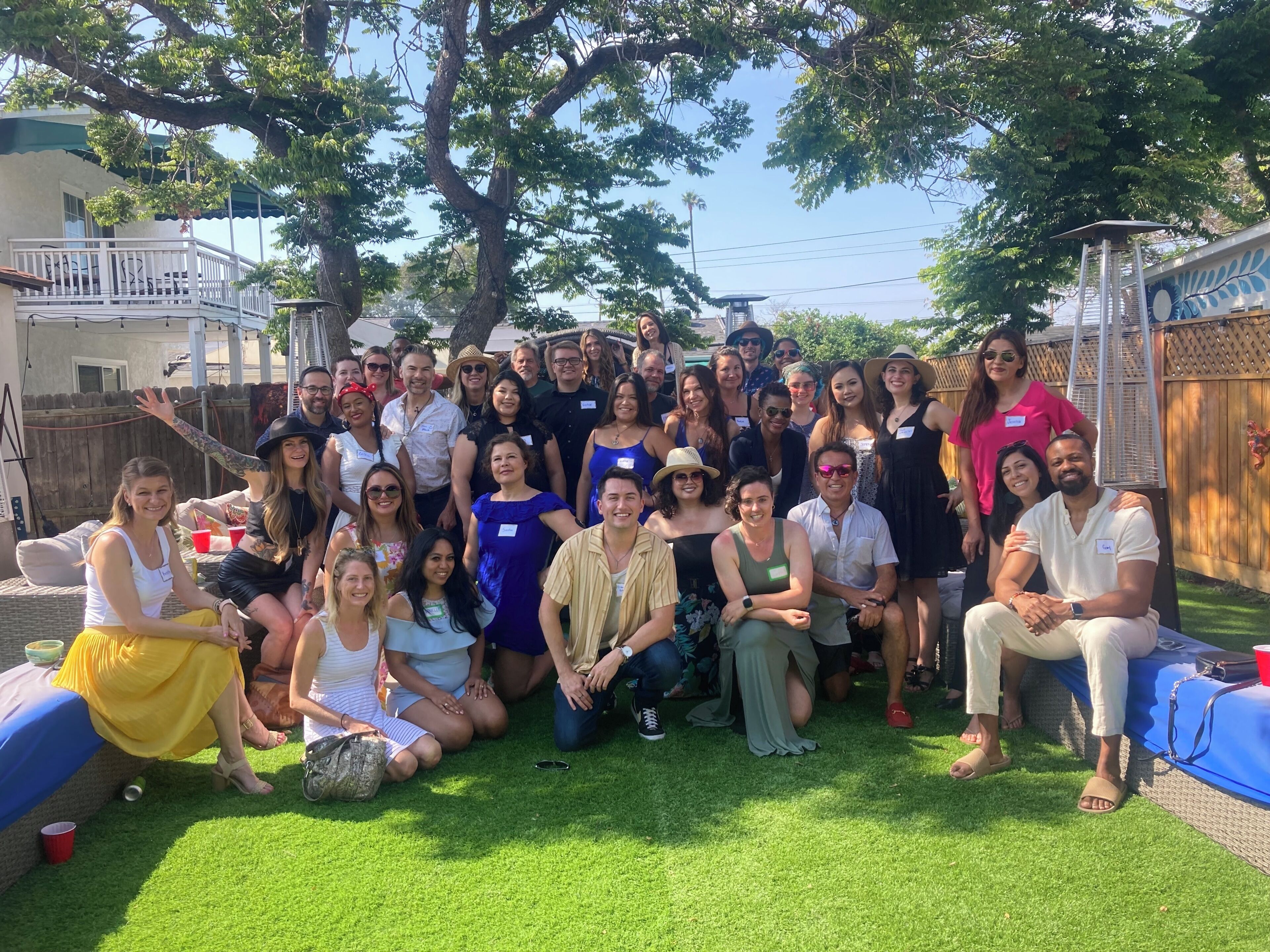 A group of people gathered outdoors, possibly at a social event, with a wooden fence and a building in the background.