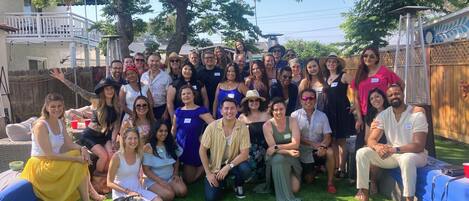 A group of people gathered outdoors, possibly at a social event, with a wooden fence and a building in the background.