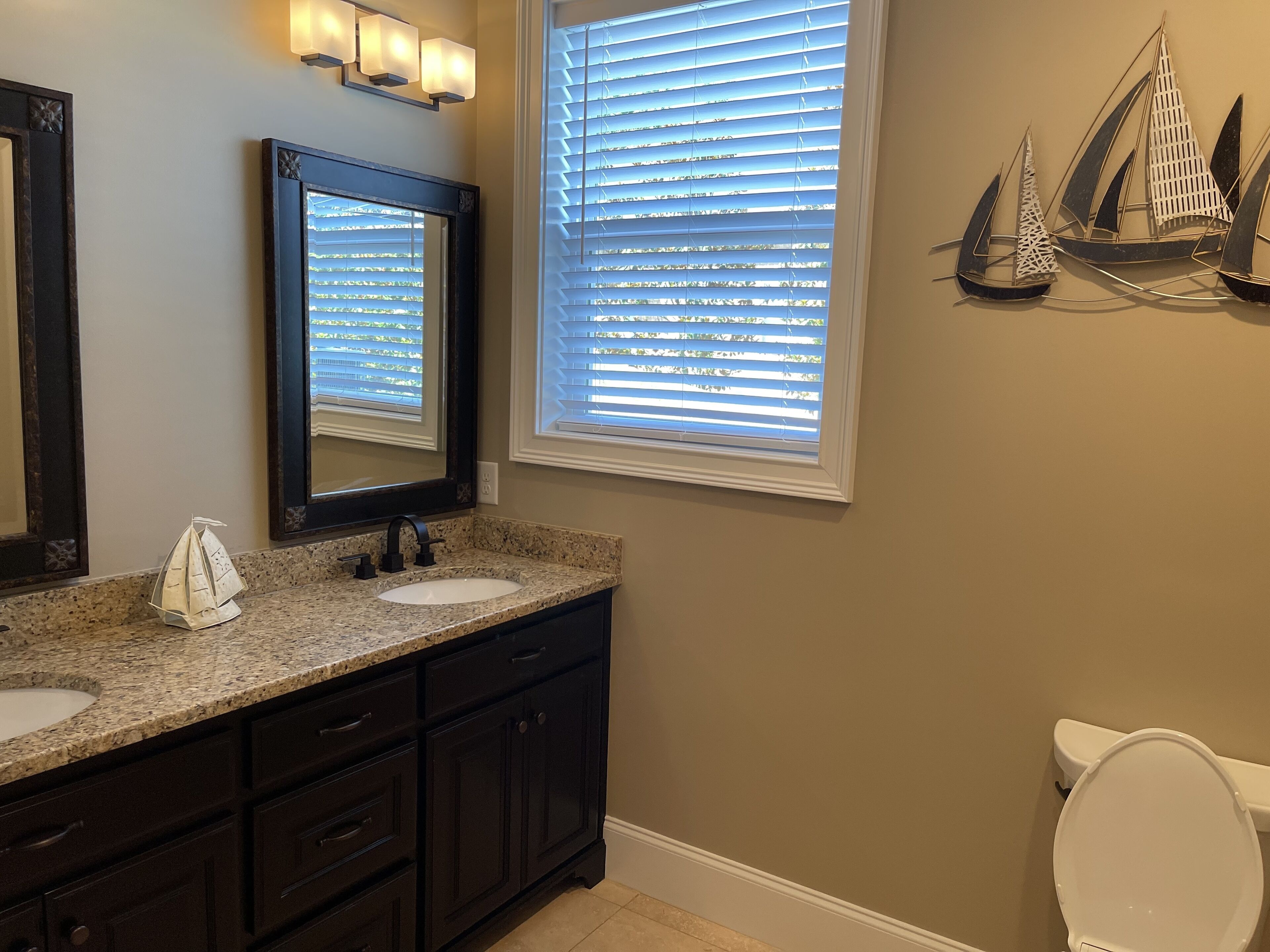 A bathroom with a dark vanity, a sink, and a toilet. There are two mirrors above the vanity, and a window with blinds.