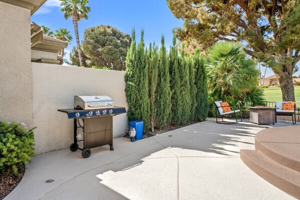 An outdoor patio with a portable grill, a seating area with a bench and table, and a backdrop of trees and a hedge.