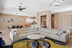A modern living room with a large L-shaped sectional sofa, a round coffee table, and a dining area with a white table and orange chairs. - Golf Course Home w/ Private Pool, Spa, Casita, Firepit & Covered Patio Dining (Palm Desert)