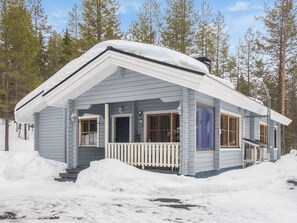 A small, single-story cabin with a porch, surrounded by snow and trees.