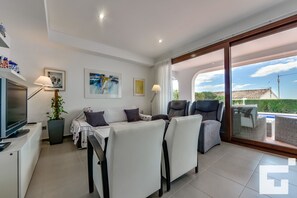 A modern living room with a sofa set, a TV, and a dining table. There is a large window with a view of a house and greenery. - Mirador de Bassetes 15 - Grupo Turis - Calpe, Costa Blanca (Calpe)
