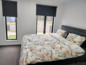 Een slaapkamer met een bloemenbedekking, twee ramen met zonwering, en uitzicht op een weg en groen buiten.