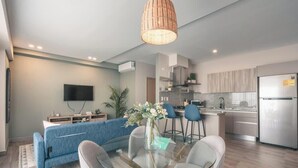Un salon moderne comprenant un canapé, une table à manger avec des chaises, un coin cuisine avec un réfrigérateur et un téléviseur fixé au mur.