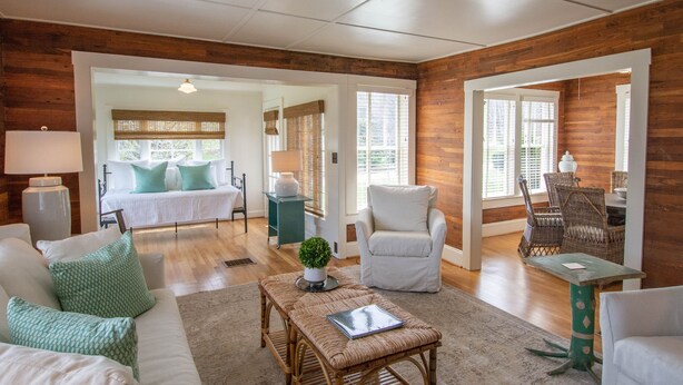 A spacious living area with a large sofa, a coffee table, and a dining table. There is a white armchair and a wicker chair. The room has wooden walls and a ceiling with a light fixture. - Homeplace 1936 | Walk to Downtown West Jefferson | Historic High Country Stay (West Jefferson)