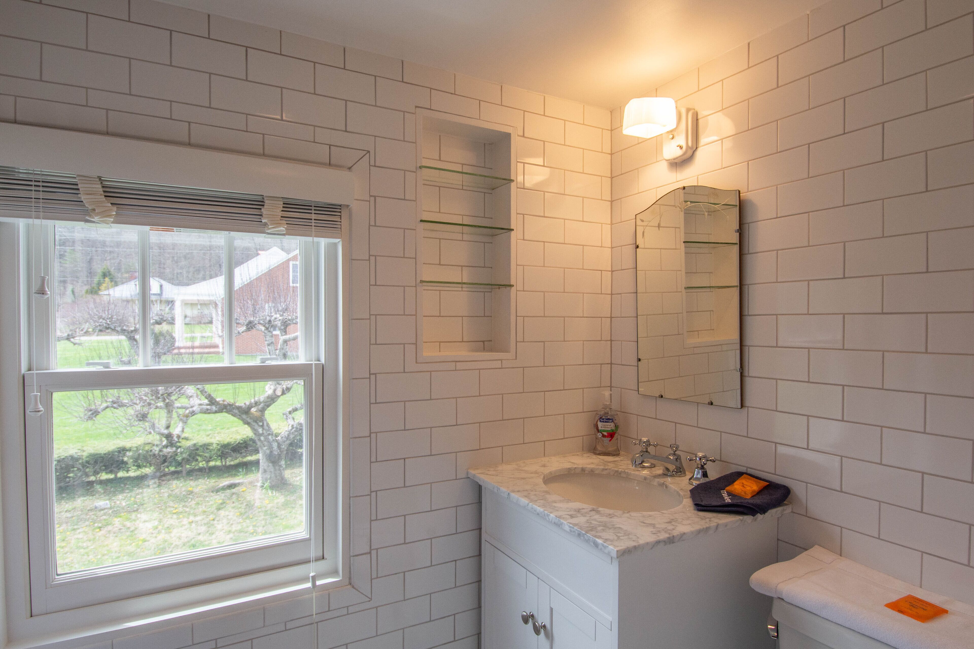 A bathroom with white tiled walls, a window, a sink, a mirror, and a towel on the counter.