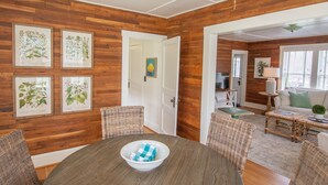 A dining area with a wooden wall, framed botanical prints, wicker chairs, a round table with a bowl, and a view into a living room with a sofa and a lamp. - Homeplace 1936 | Walk to Downtown West Jefferson | Historic High Country Stay (West Jefferson)