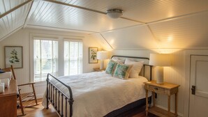 A bedroom with a wooden bed, bedside lamps, a rocking chair, and a window with blinds. - Homeplace 1936 | Walk to Downtown West Jefferson | Historic High Country Stay (West Jefferson)