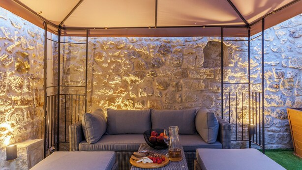 Un rincón al aire libre acogedor con un muro de piedra, un banco acolchado y una mesa con aperitivos y bebidas.