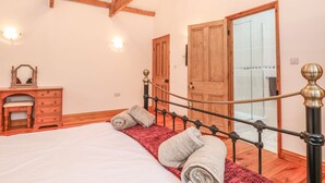 A bedroom with a wooden bed frame, a red throw blanket, and two towels. There is a wooden dresser and a mirror on the left. The room has wooden beams on the ceiling and a door leading to a bathroom with a glass shower. - Coastal Barn Conversion, overlooking the stunning Cot valley. (Saint Just)