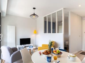 A modern living room with a flat-screen TV, a dining table set with breakfast, and a large window with a view.