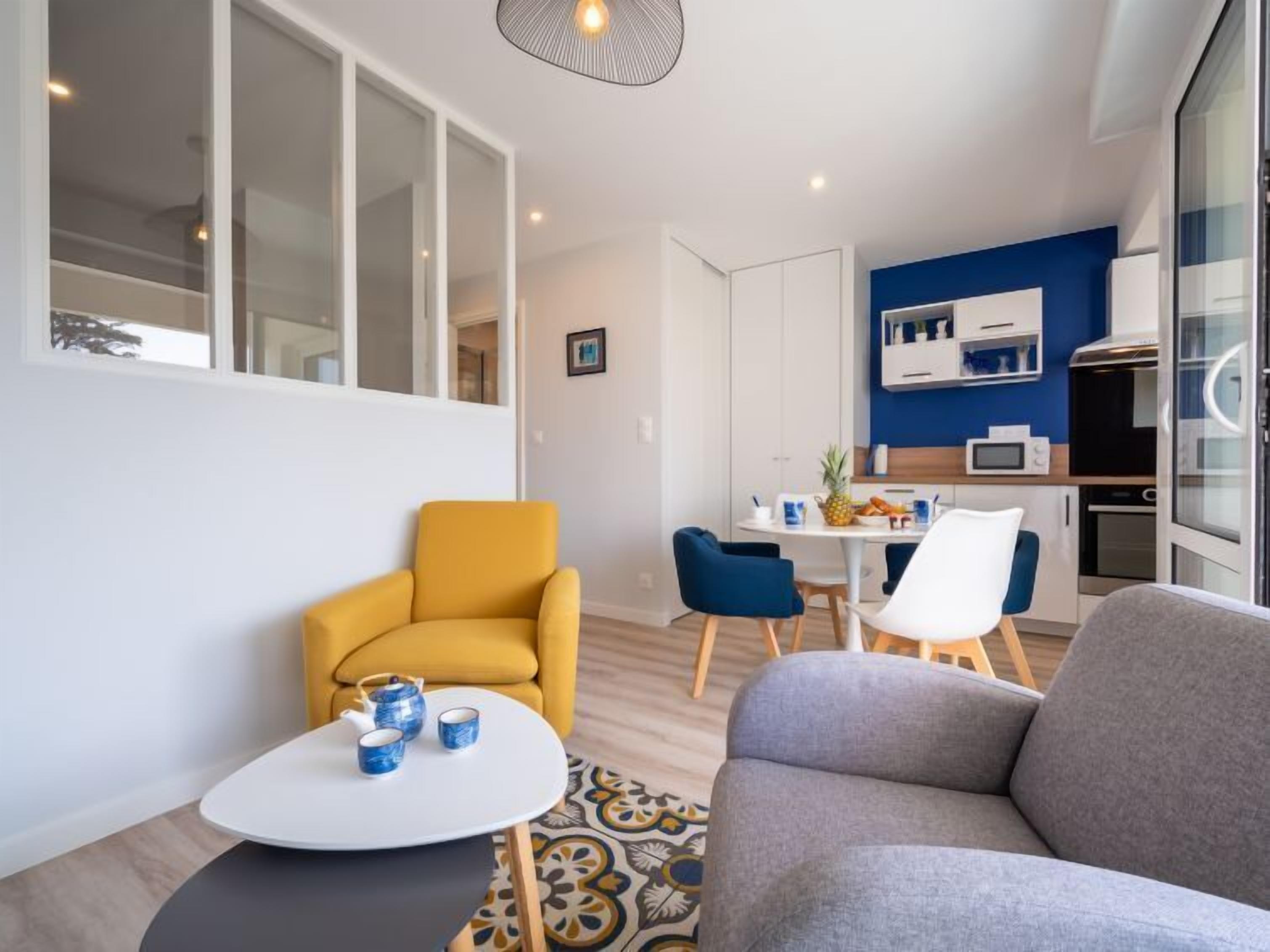 Una sala moderna con sofá, un sillón amarillo y una pequeña mesa de centro. Se observa un comedor con una mesa blanca y sillas azules. Se puede ver la cocina, con armarios blancos y una pared azul de acento.