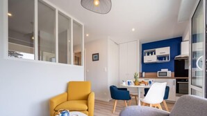 En moderne stue med en sofa, en gul lænestol og et lille sofabord. Der er et spiseområde med et hvidt bord og blå stole. Køkkenet er synligt med hvide skabe og en blå accentvæg.