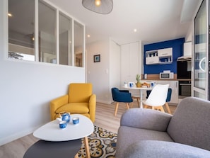 A modern living room with a sofa, a yellow armchair, and a small coffee table. There is a dining area with a white table and blue chairs. The kitchen is visible with white cabinets and a blue accent wall.