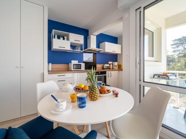 A modern kitchen with white cabinets, a microwave, and a coffee maker. There is a dining table with a white tablecloth, a bowl of fruit, and a pineapple. - Port An Dro by Interhome (Carnac)
