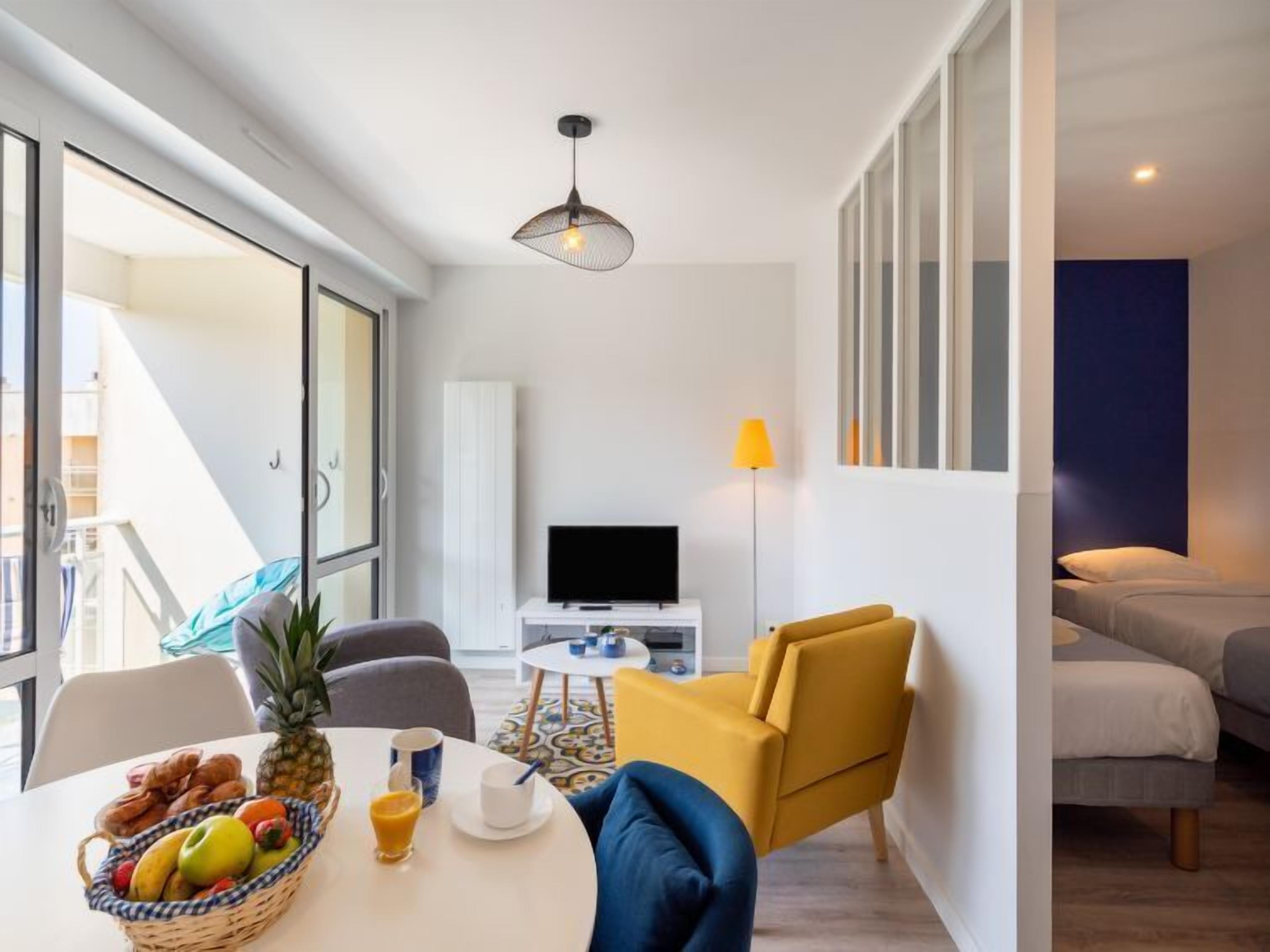 Een moderne woonkamer met een televisie, een eettafel met fruit en drankjes, een bank en een gele fauteuil.
