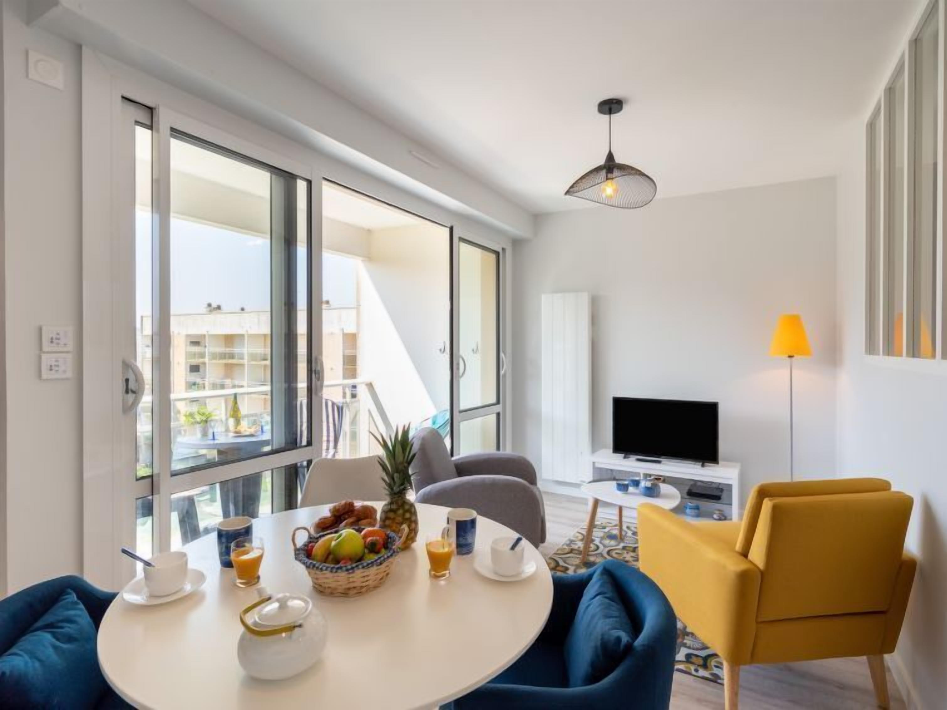 A modern living room with a round table, blue chairs, a TV, and a balcony with a view of other buildings.