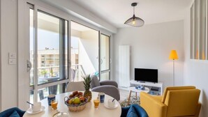 En moderne stue med et rundt bord, blå stole, et fjernsyn og en balkon med udsigt til andre bygninger.