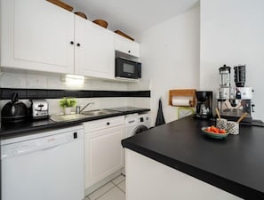Une cuisine moderne avec des meubles blancs, un plan de travail noir, un four à micro-ondes, une machine à café et un égouttoir à vaisselle contenant un bol de fruits.