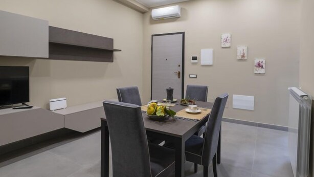 A modern dining area with a dark wooden table, four grey chairs, and a fruit bowl on the table. There is a flat-screen TV on a white stand to the left. The room has a neutral color scheme with beige walls and a grey floor. - Daydreaming by Interhome (Meta di Sorrento)