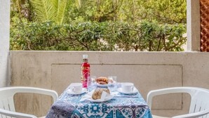 Ein Tisch draußen mit einer gemusterten Tischdecke, zwei weiße Stühle, eine Wasserflasche, ein Teller mit Essen und eine Tasse.