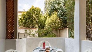 Un petit espace extérieur aménagé pour prendre les repas, avec une table pour deux, entouré de chaises blanches, et une vue sur des arbres et un bâtiment en arrière-plan.