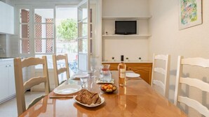 Un coin repas avec une table en bois, des chaises blanches, un téléviseur fixé au mur et une fenêtre donnant sur un paysage verdoyant.