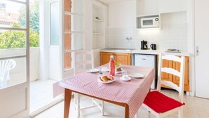 Eine helle Küche mit weißen Schränken, einer Mikrowelle und einer Kaffeemaschine. Es gibt einen Esstisch mit einer rosa Tischdecke und einem roten Stuhl.