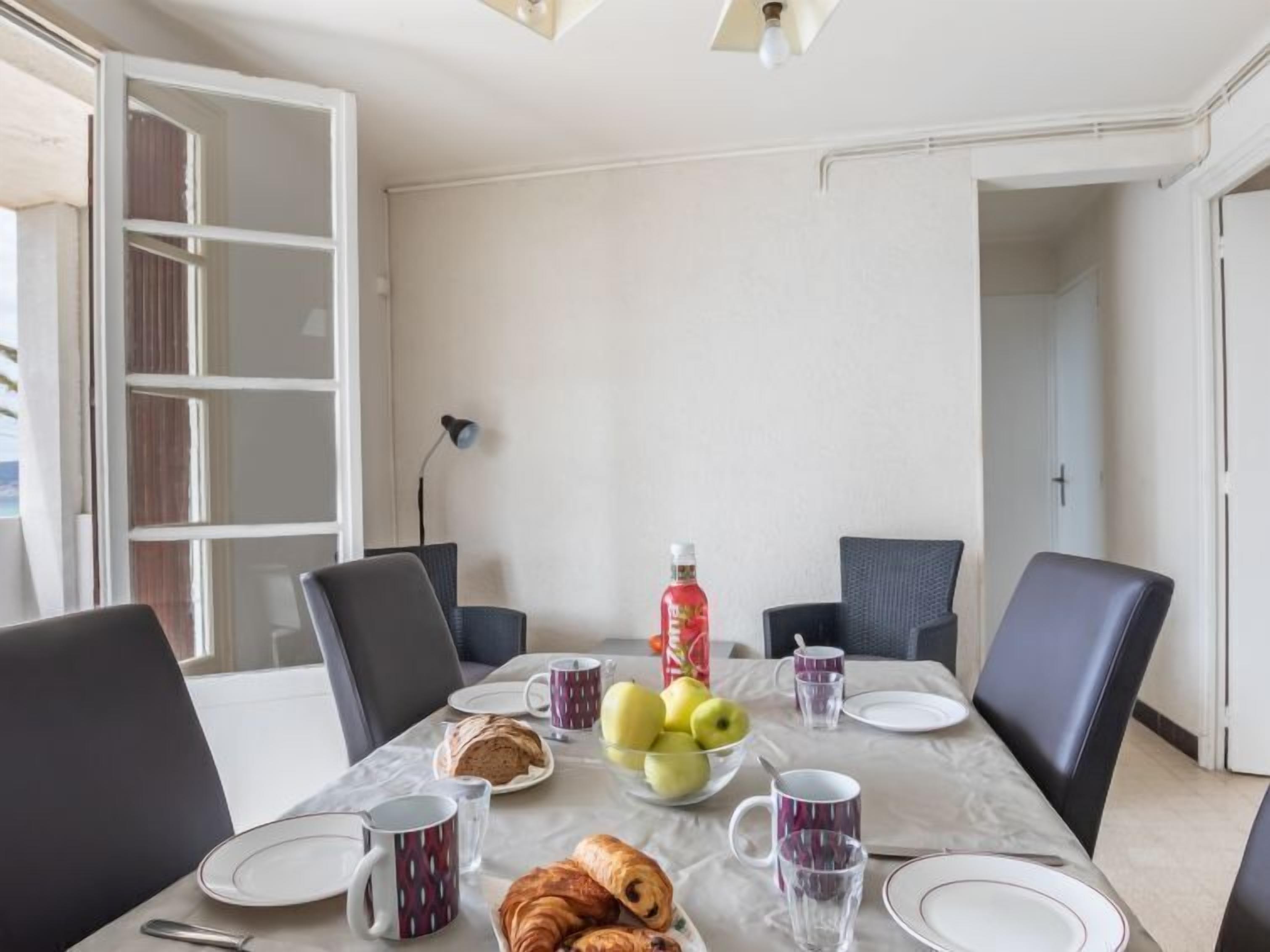 Un coin repas avec une table dressée pour quatre personnes, comprenant des assiettes, des couverts et une variété d’aliments, comme du pain, des pommes et des croissants.