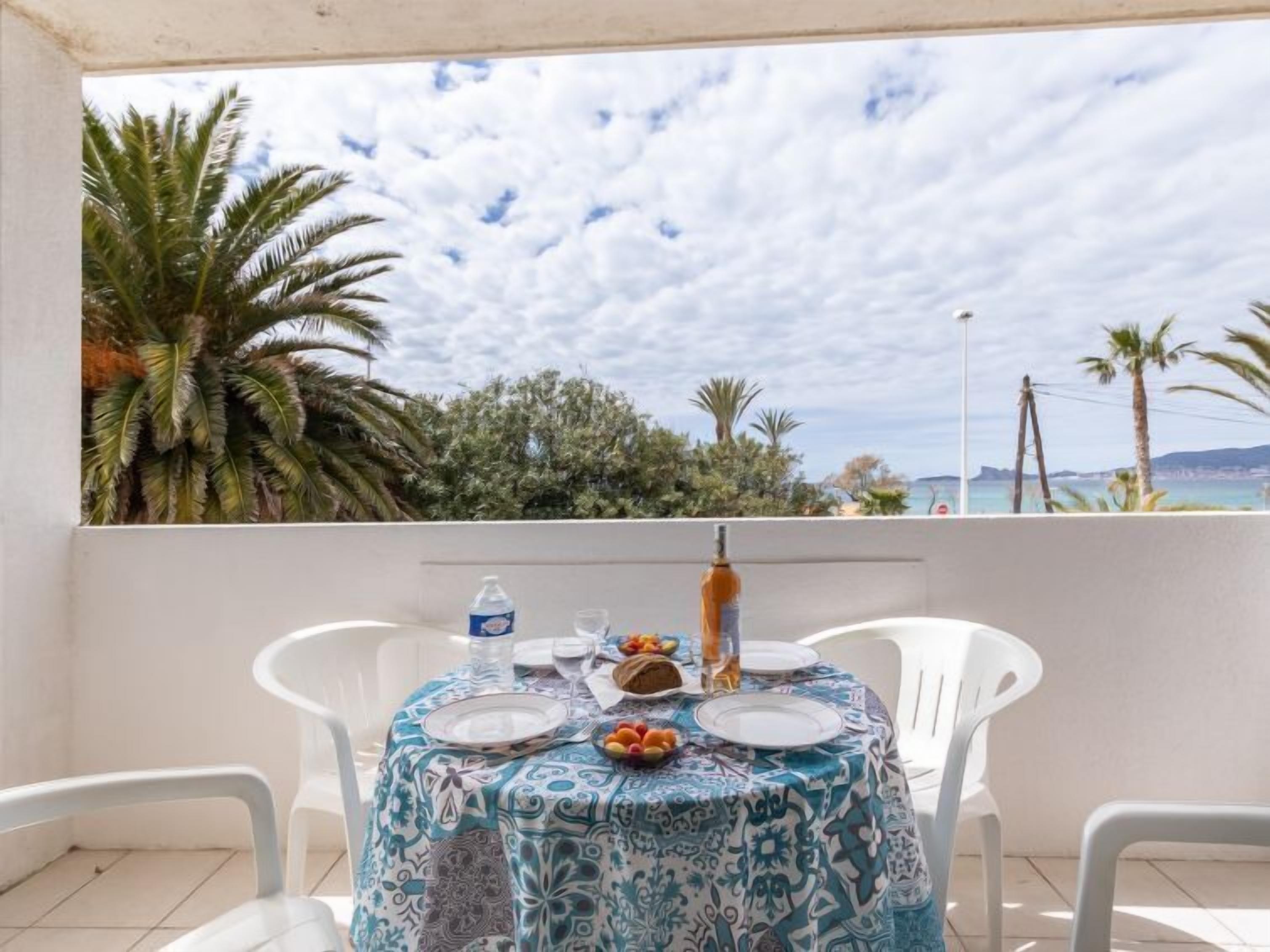 A table set for two with a patterned tablecloth, white chairs, and a view of the sea and palm trees.