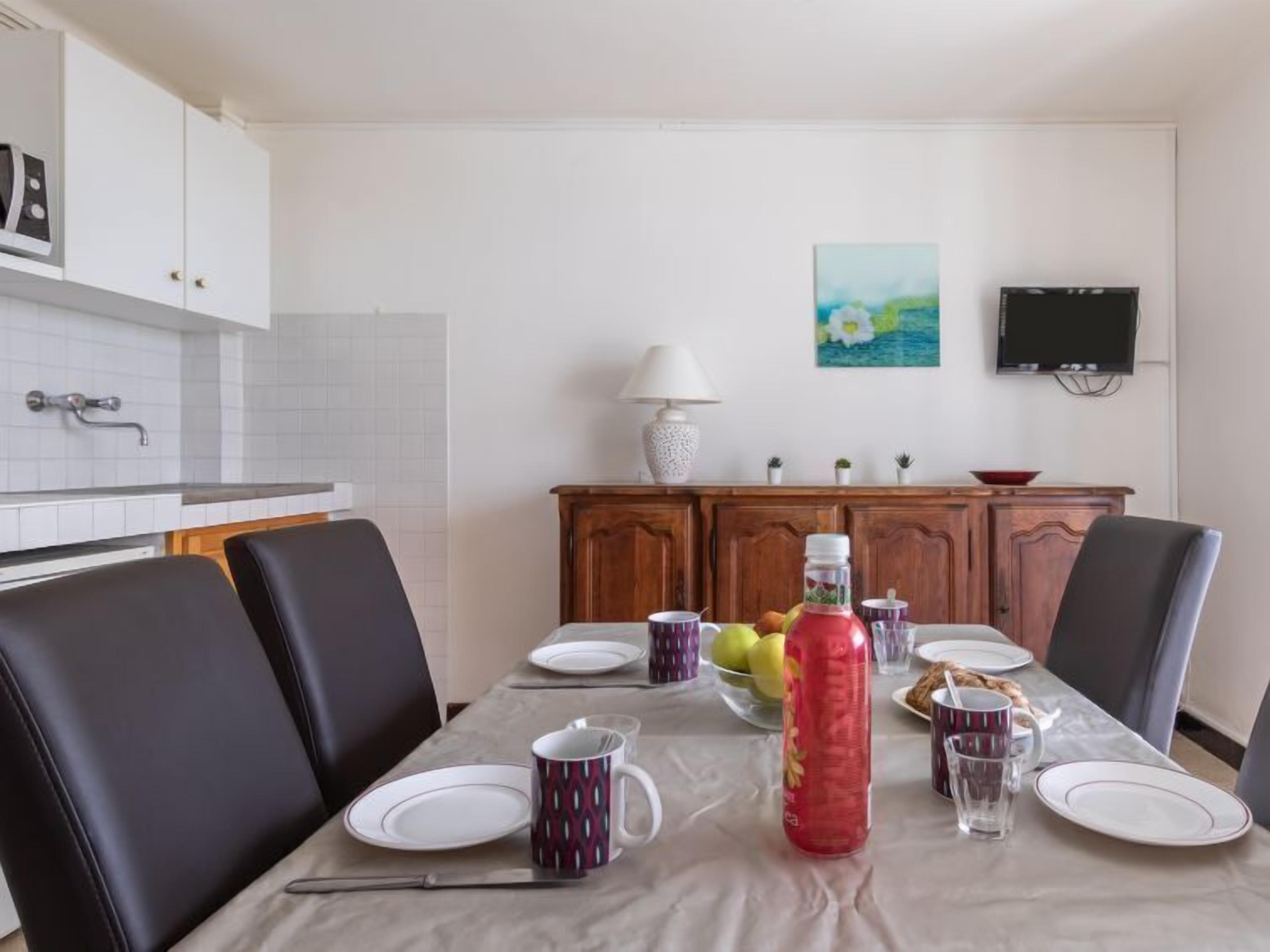 Ein Essbereich mit einem Tisch, der für vier Personen gedeckt ist, inklusive Tellern, Tassen und einer Flasche. Um den Tisch herum befinden sich Stühle, und an der Wand ist ein Fernseher angebracht.