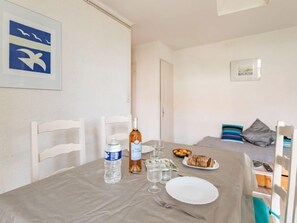 Mesa de jantar posta com garrafa, copos, prato e garfo. Há uma cadeira com almofada branca e um travesseiro listrado. Uma porta é visível ao fundo.