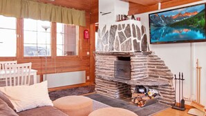 Ein gemütliches Blockhaus-Interieur mit Kamin aus Stein, einem an der Wand montierten Fernseher und einem Holzespeisesaal mit Stühlen.
