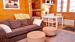 Ein gemütliches Blockhaus-Interieur mit einem braunen Sofa, zwei Hocker, einem Esstisch mit Stühlen, einer Lampe und einem gerahmten Bild an der Wand.