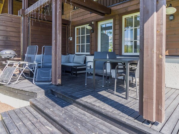 Een houten terras met buitenmeubilair, waaronder stoelen en een bank, en een barbecue.