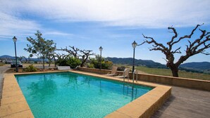 Une piscine rectangulaire à l’eau d’un bleu limpide, entourée d’une terrasse en bois et d’un petit muret en pierre. Des chaises longues et un banc sont disposés le long du bord de la piscine, et des arbres ainsi qu’un lampadaire se trouvent à proximité.